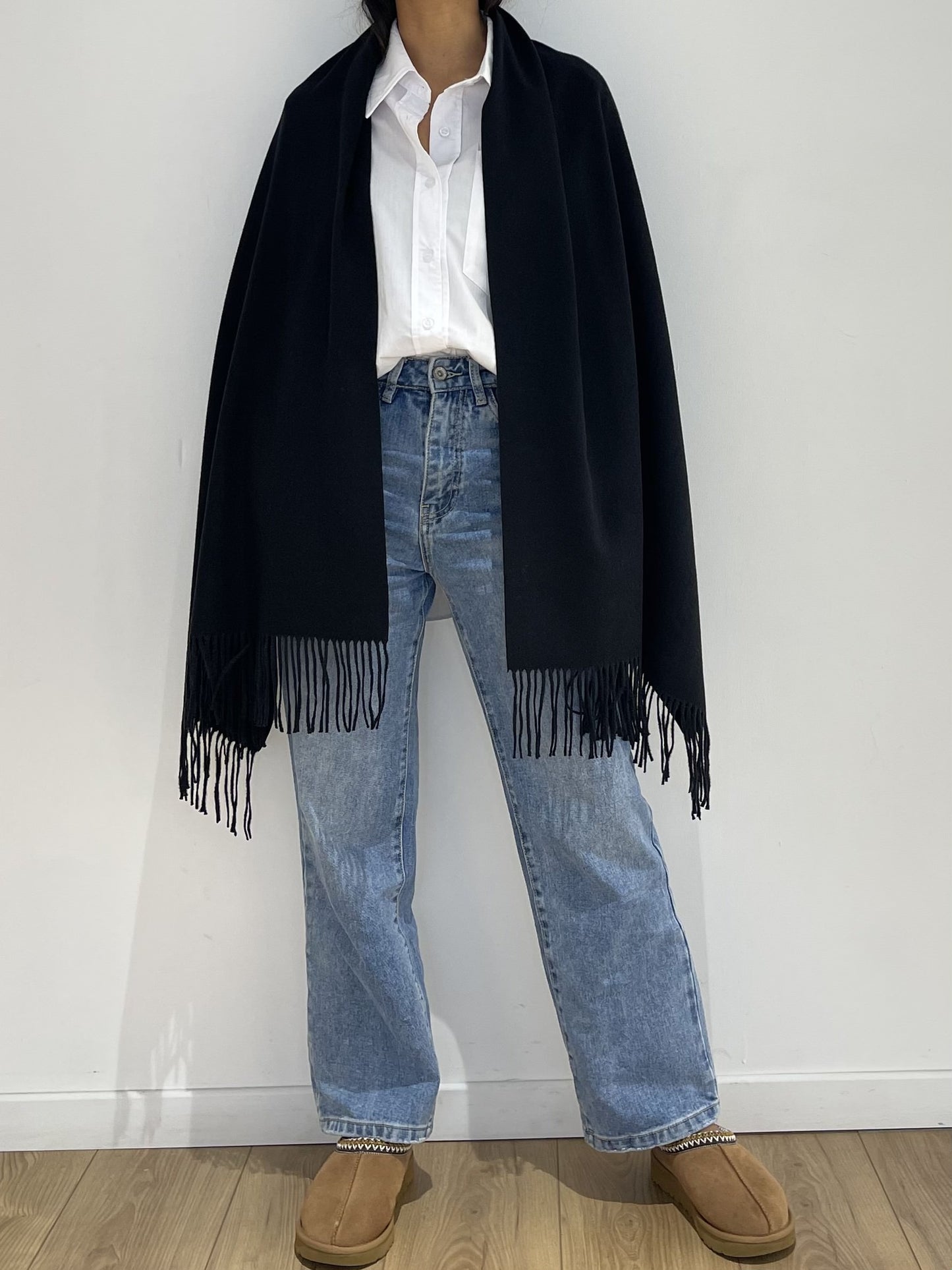Étole à franges façon poncho en noir, enveloppant les épaules pour un look sophistiqué, assorti à un jean et des chaussures en daim.
