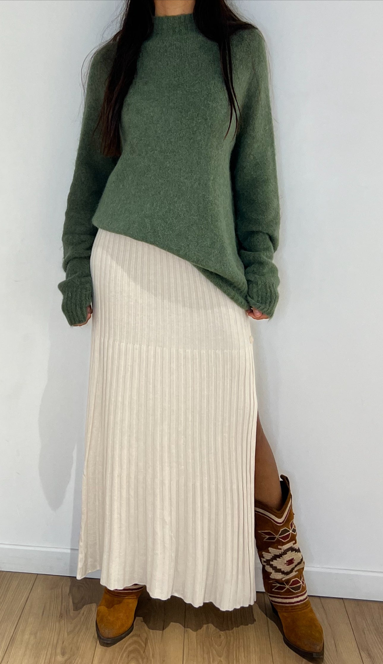 Jupe longue plissée fendue en écru associée à un pull vert doux et des bottes cowboy, parfaite pour un look bohème chic.
