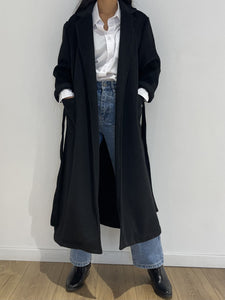 Femme élégante portant un Manteau noir long de Kata Paris, parfaitement assorti avec une chemise blanche et un jean bleu pour un style classique et chic.