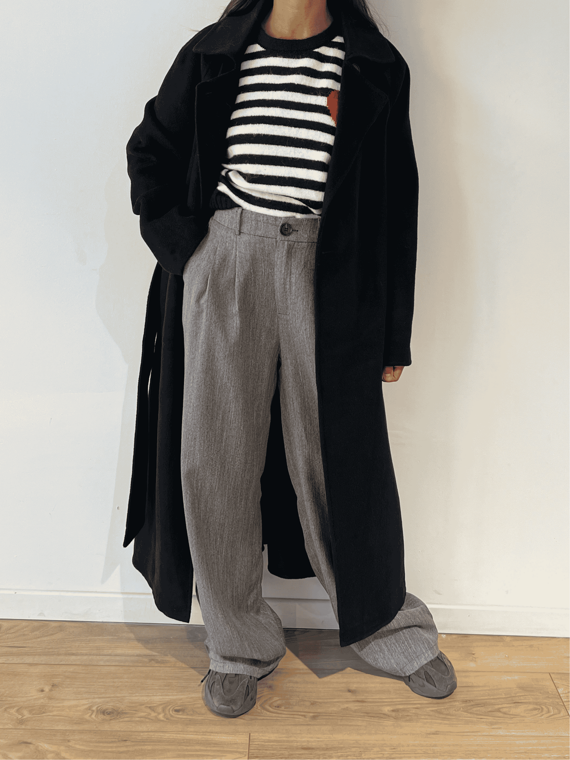 Femme portant un pantalon gris ample avec un manteau noir et un pull à rayures, prise de vue de face.