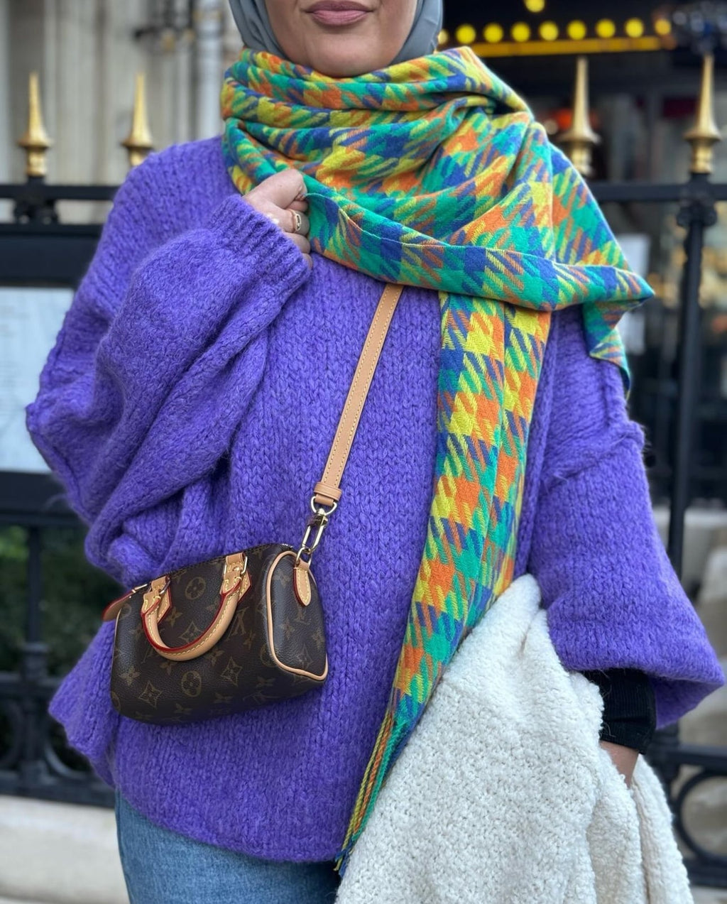 Écharpe XXL en laine multicolore dans les tons jaune, orange et vert, portée avec un pull violet et un sac de marque.