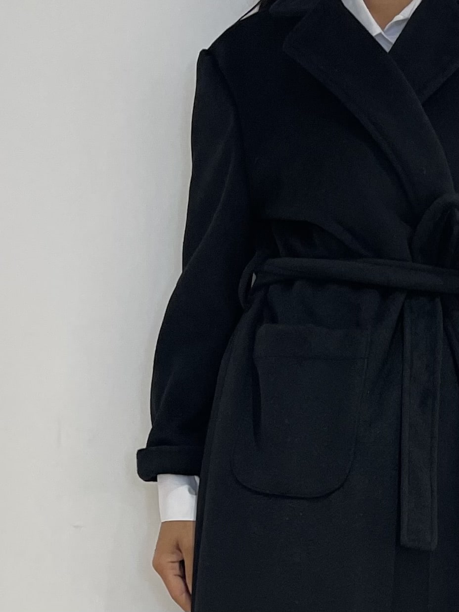 Zoom sur le Manteau noir ceinturé de Kata Paris, montrant les détails de sa coupe impeccable et sa texture de qualité.